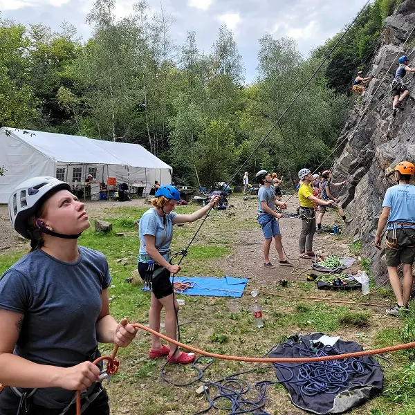 Een groep mensen met helmen en klimgordels is buiten aan het rotsklimmen. Sommigen beklimmen de rotswand, terwijl anderen hen zekeren of toezicht houden. Op de achtergrond staan een witte tent en bomen. Klimuitrusting ligt verspreid op de grond.